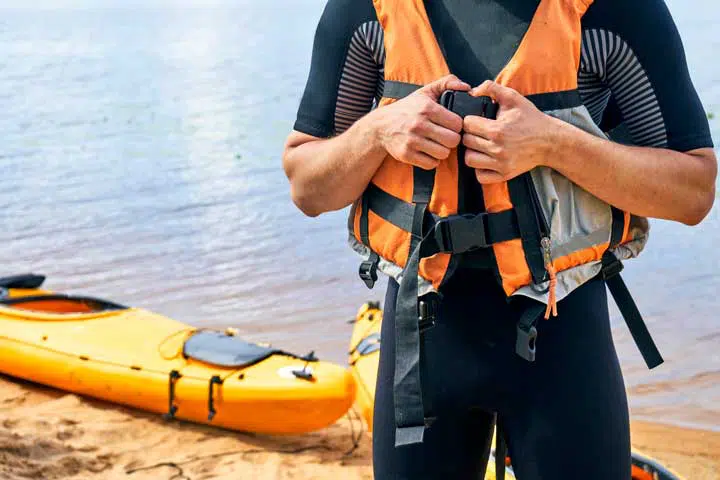 Young male hiker wearing wetsuit putting on a life vest with a yellow kayak. 